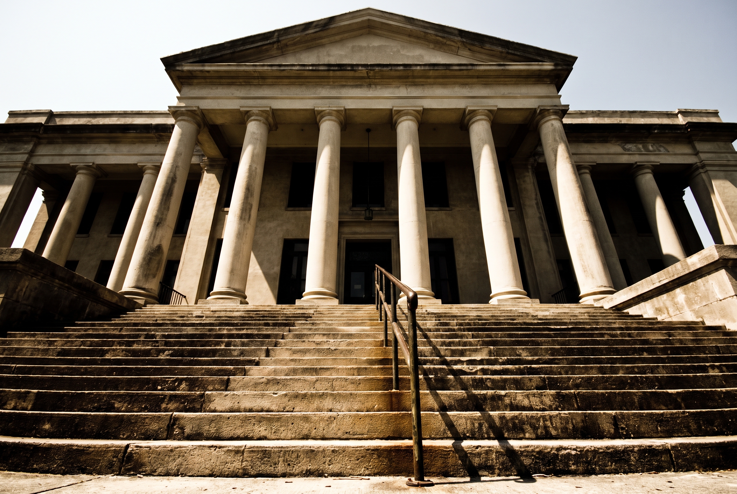 A steep, empty stone staircase rises toward fluted courthouse columns and a plain pediment under harsh midday sun.