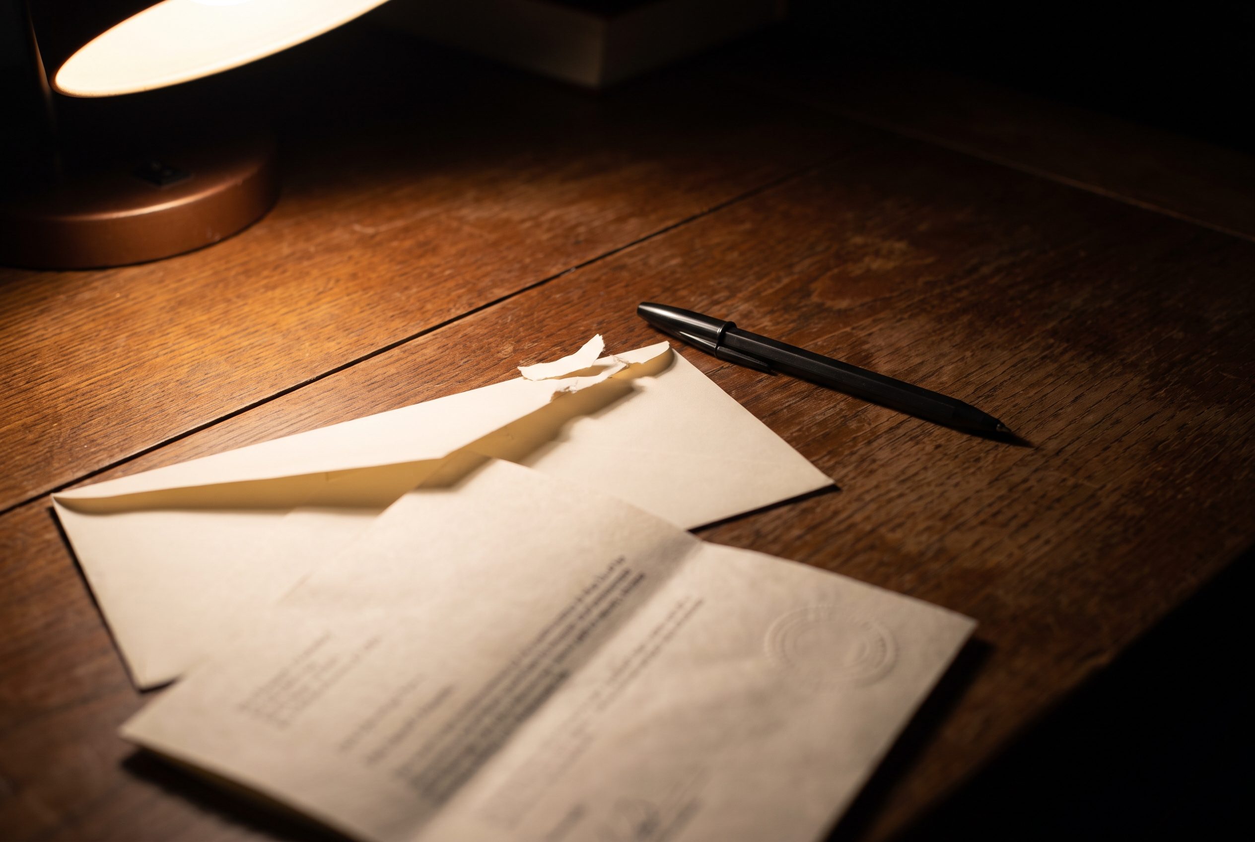 A letter pulled halfway from its envelope on a dark wood desk, lit by a single lamp, with a pen resting nearby.
