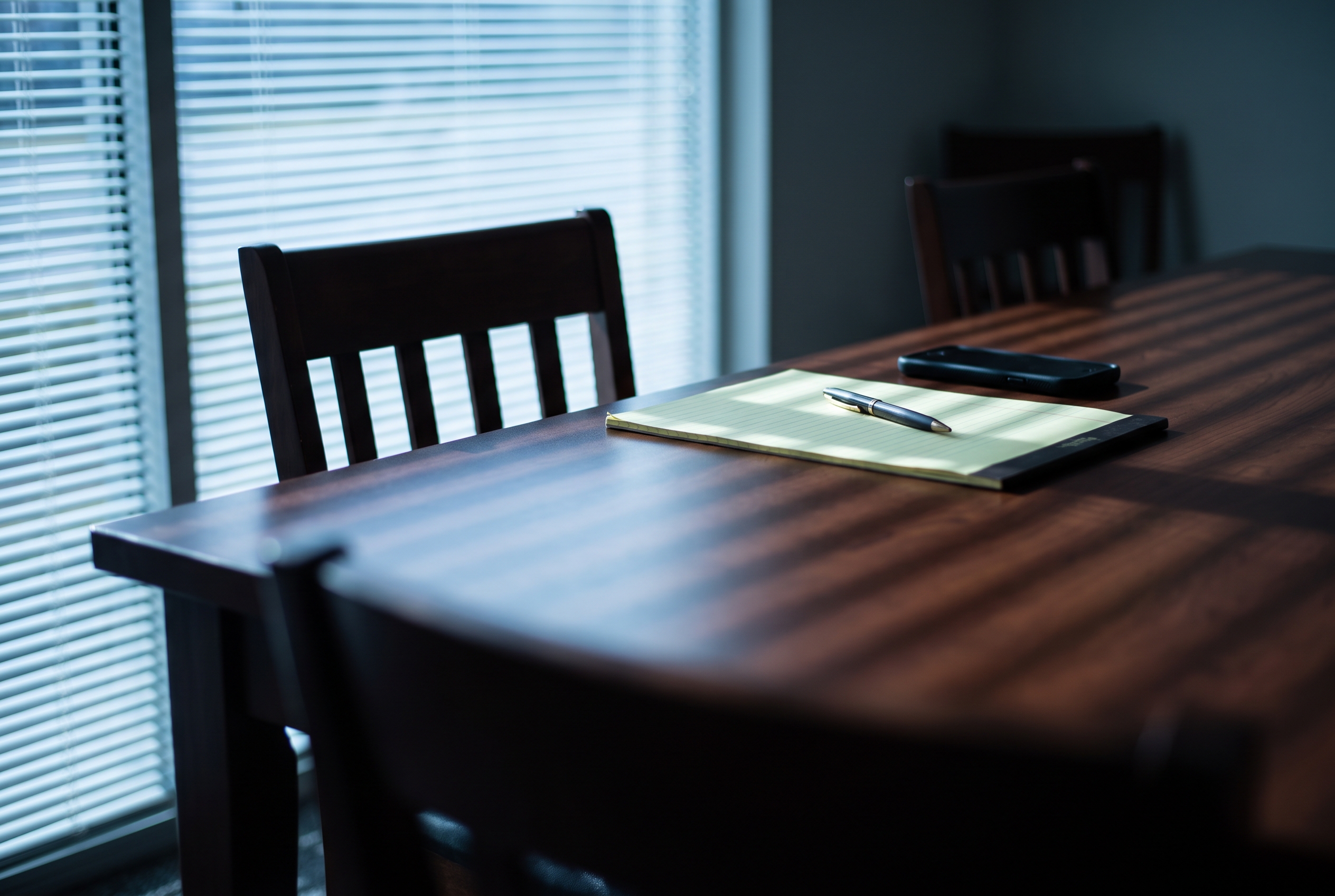 An empty wood conference table holding a legal pad, pen, and a face-down phone, crossed by horizontal shadow bars from closed window blinds.