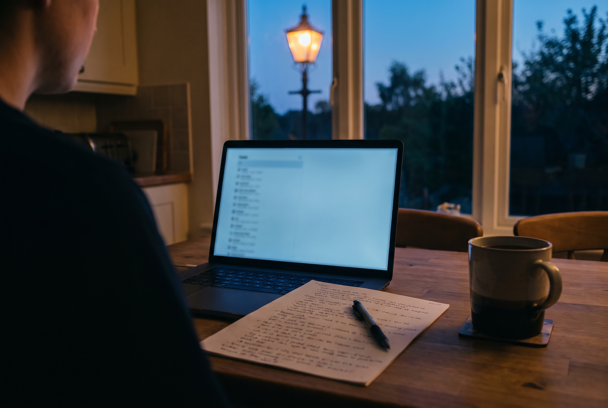 A laptop with a blurred case list glowing on a kitchen table at dusk beside handwritten notes, a pen, and a cooling mug of coffee.