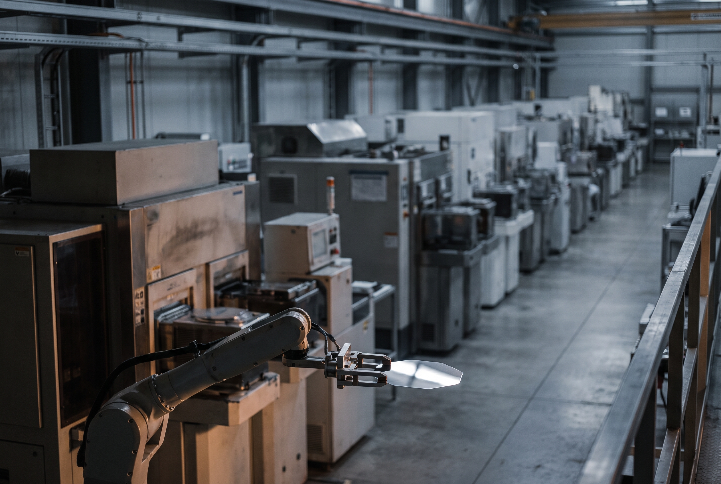 Elevated view of a wafer fabrication line with a robotic arm holding a thin hexagonal silicon carbide wafer in sharp focus against a blurred row of tools.