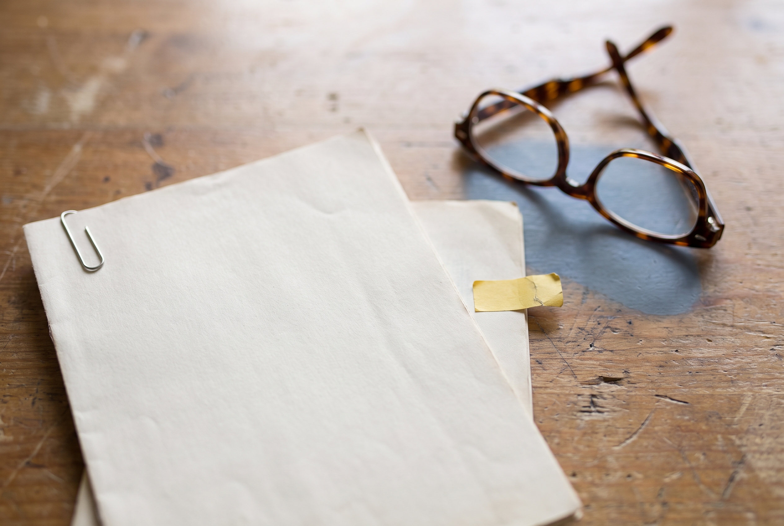 Two stacked paper documents with a paperclip and sticky tab beside a pair of open reading glasses on a wood table