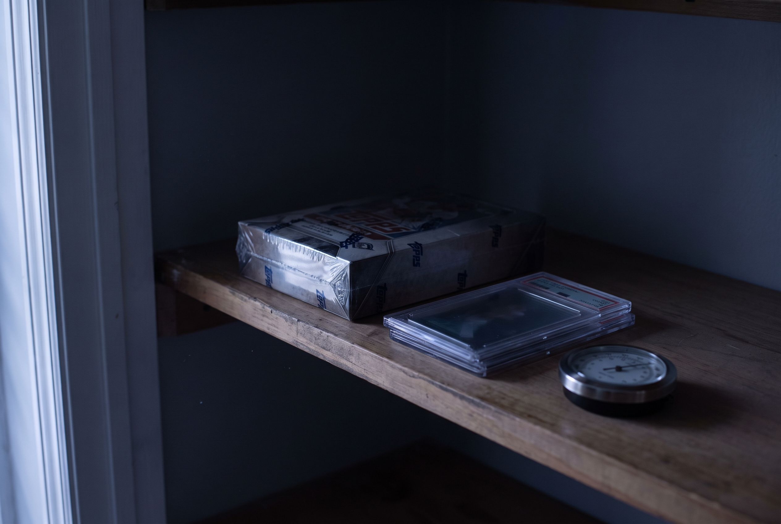 A sealed box and two graded card slabs resting on a dim closet shelf beside a small hygrometer.
